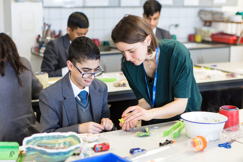 An artist educator demonstrates an activity to a pupil at a desk.