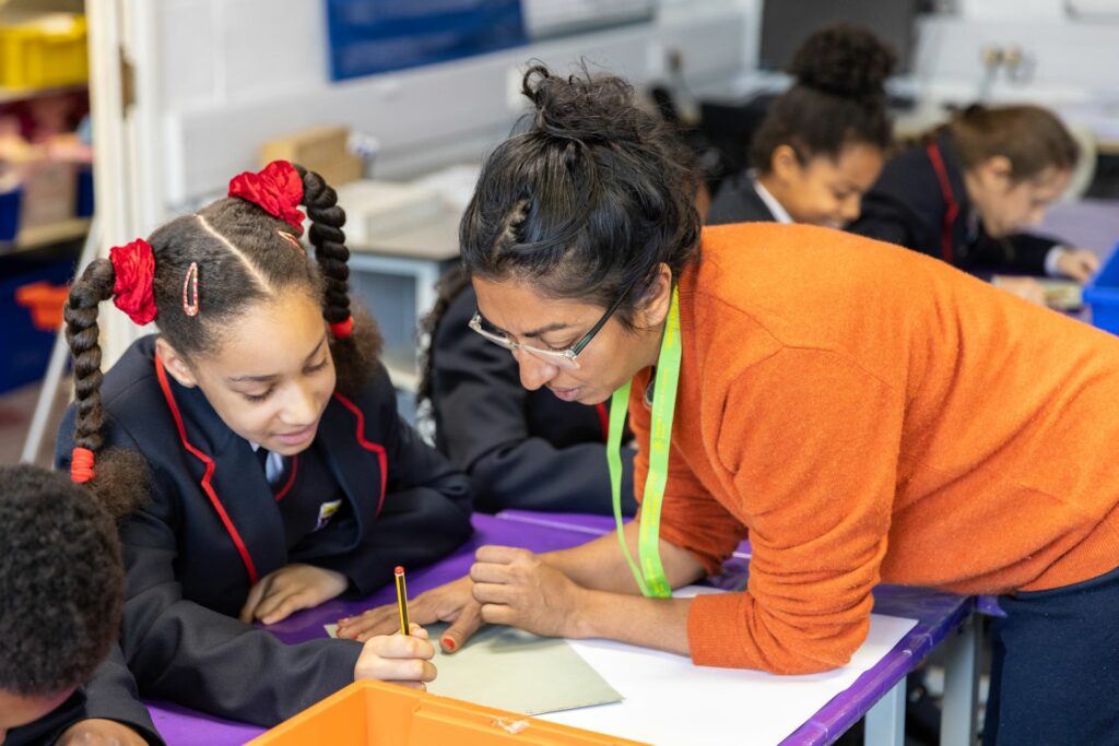 Children draw at the desks, in the forefront of the image an artist educator leans over to look at a pupil's work