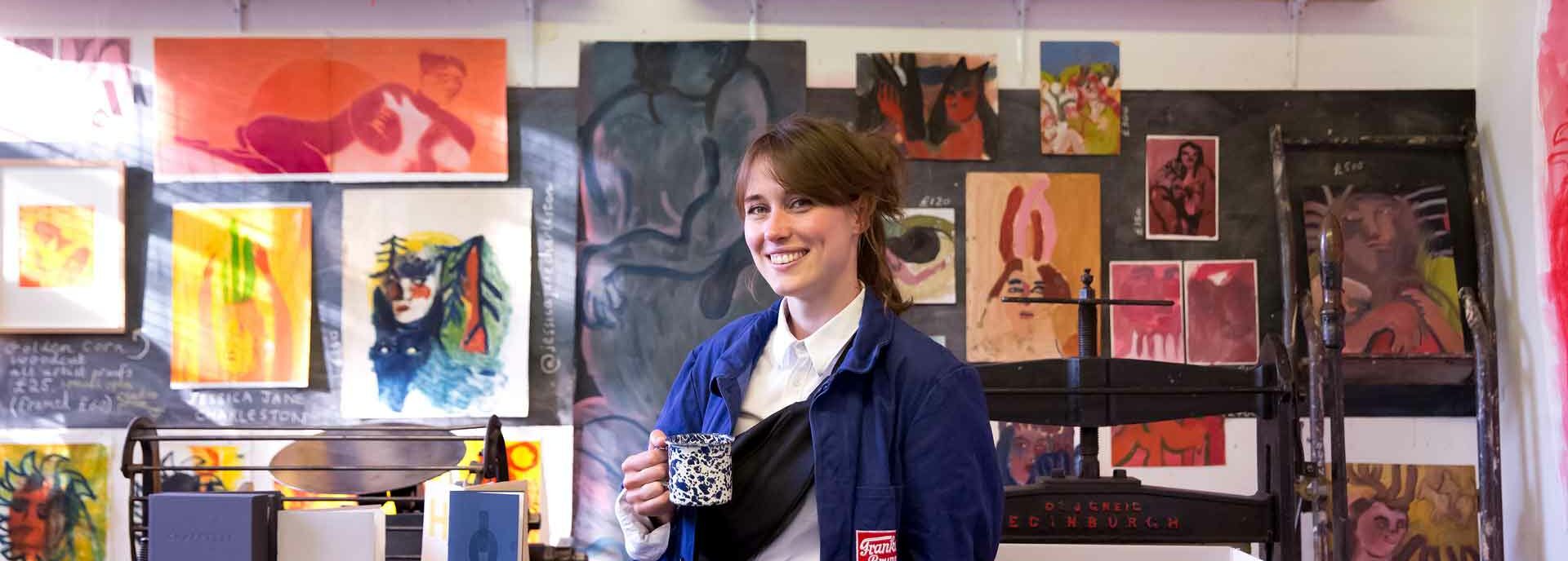 An image of an artist in their studio holding a cup of tea, with paintings behind them