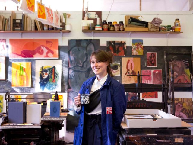 An image of an artist in their studio holding a cup of tea, with paintings behind them
