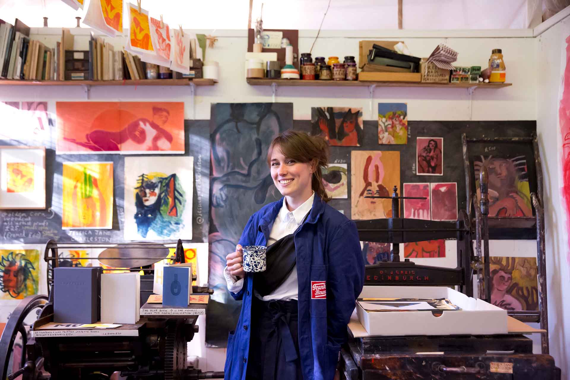 An image of an artist in their studio holding a cup of tea, with paintings behind them