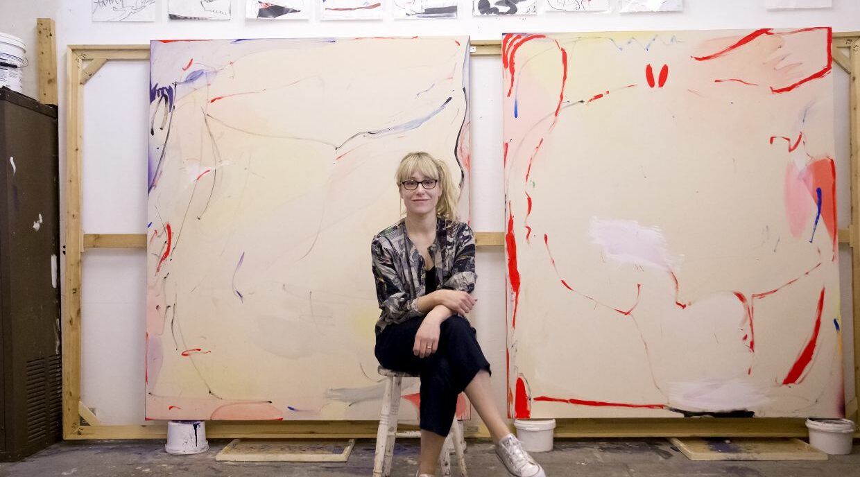 An artist sits in their studio with canvases behind them