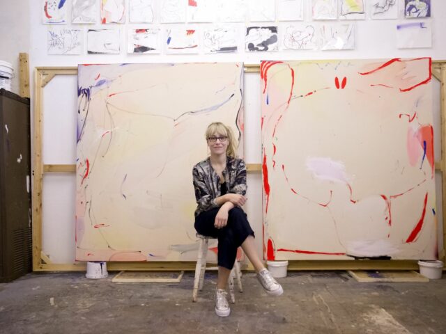 An artist sits in their studio with canvases behind them