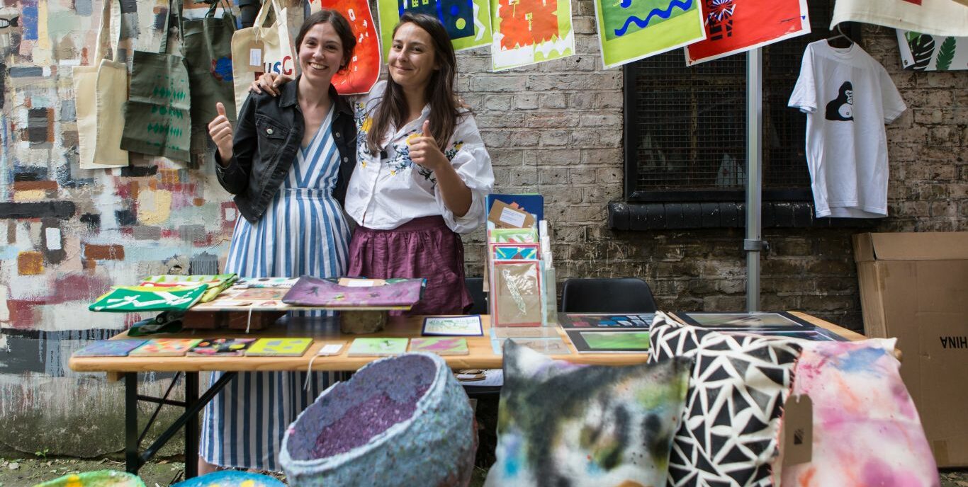 Two artists put thumbs up behind their market stall selling cushions, paintings, prints and cards
