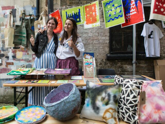 Two artists put thumbs up behind their market stall selling cushions, paintings, prints and cards