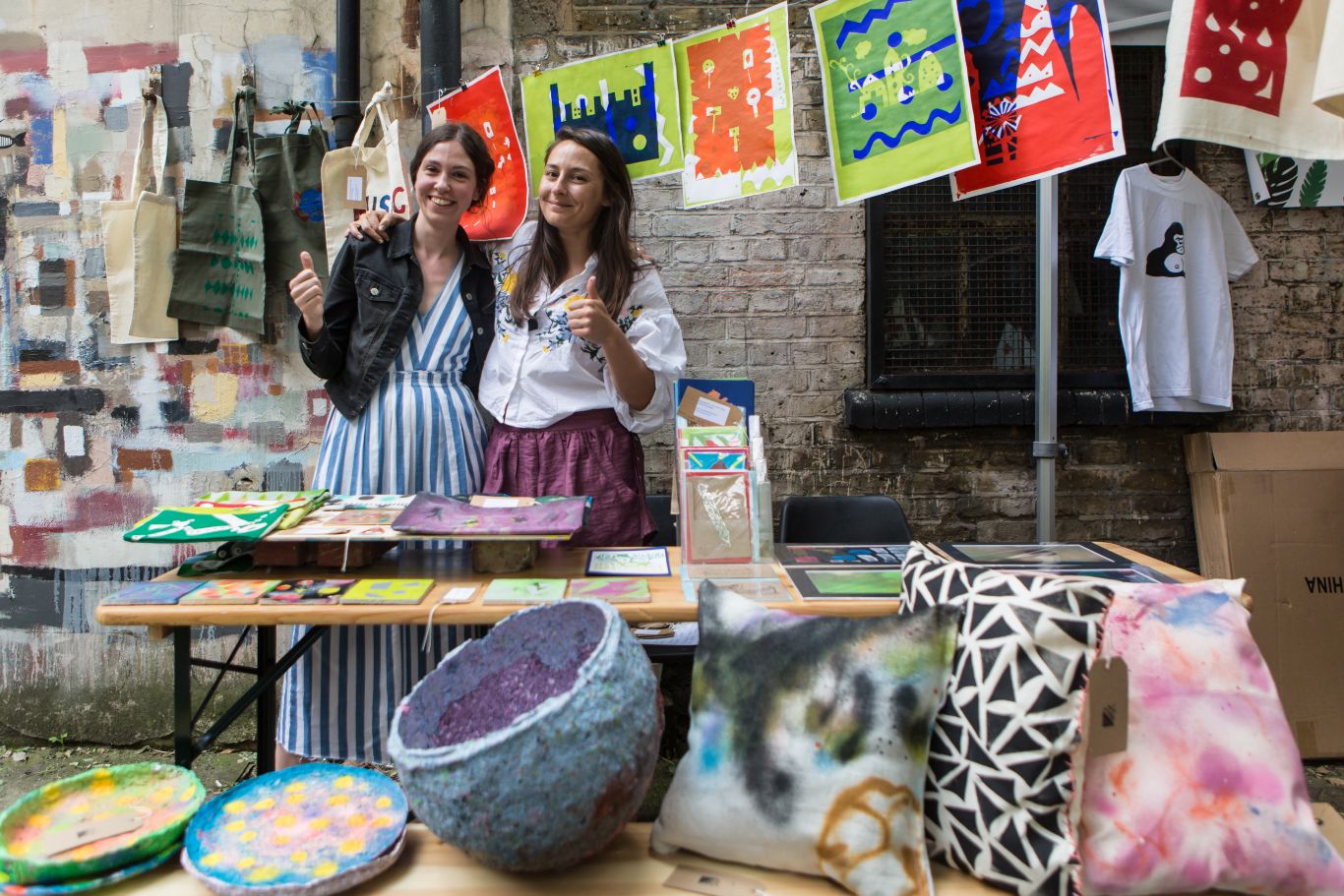 Two artists put thumbs up behind their market stall selling cushions, paintings, prints and cards