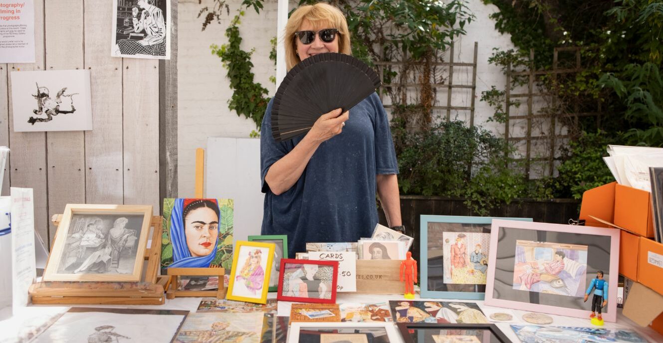 An artist behind their stall holds a fan to their face, drawings sit on the table
