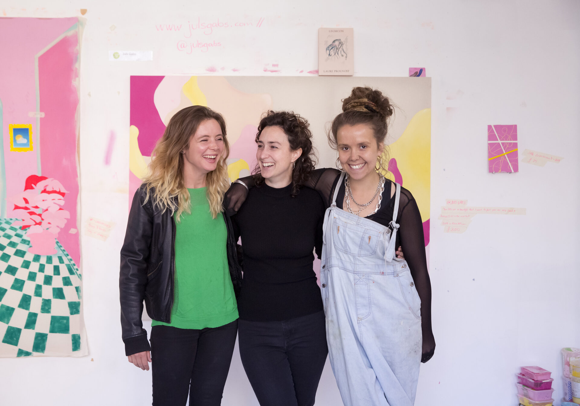 Three people loop arms smiling in a studio with a painting behind them