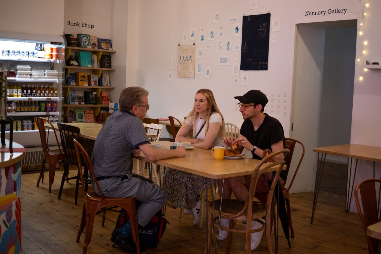 Three people sit at a table drinking coffee and eating pastries, a door behind them is open labelled Nunnery Gallery