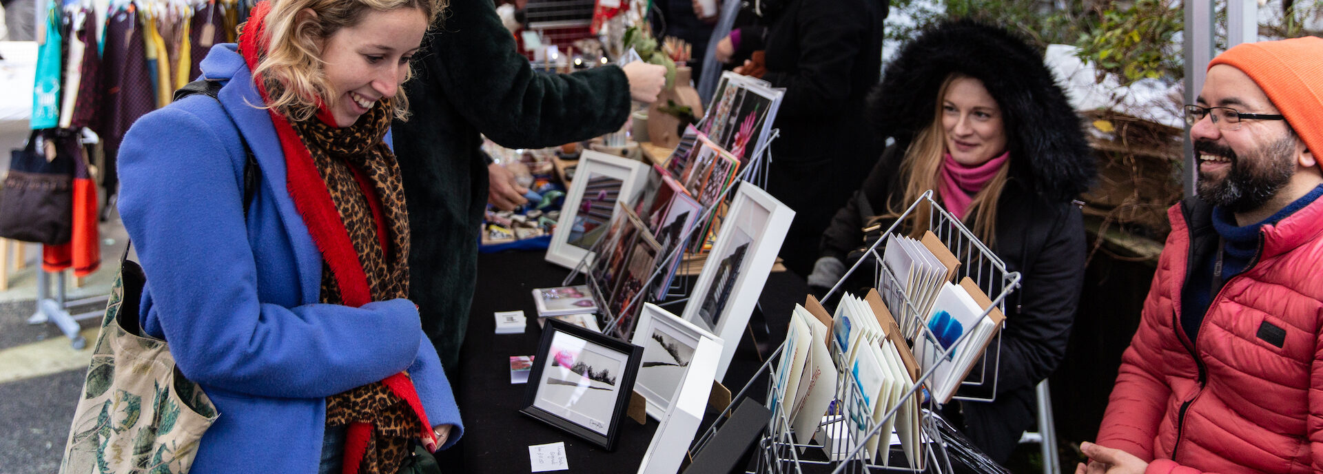 A person looks at greeting cards at a market stall, chatting to the two sellers across the table