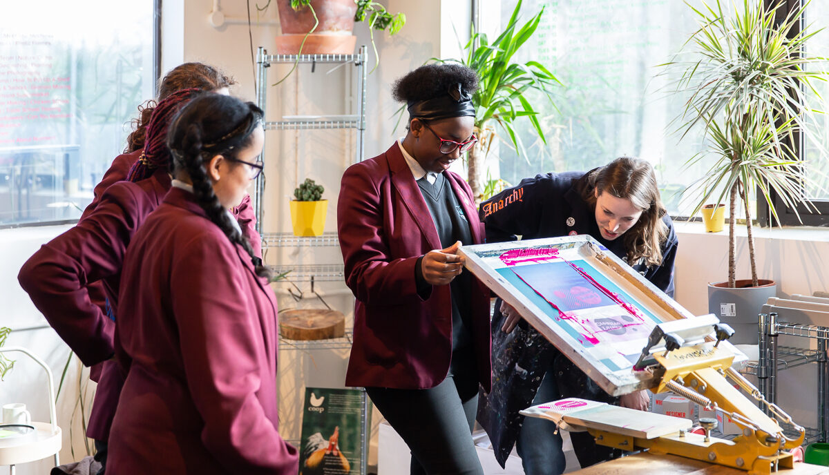 A student lifts a printmaking press helped by an artist educator, watched by students