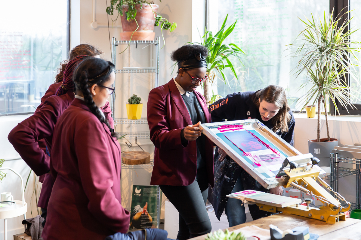 A student lifts a printmaking press helped by an artist educator, watched by students