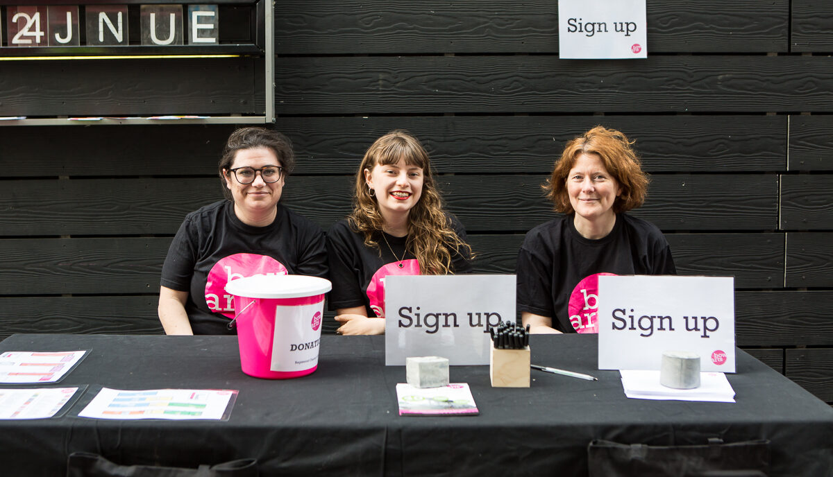 Three Volunteers sit at a welcome desk smiling at the camera
