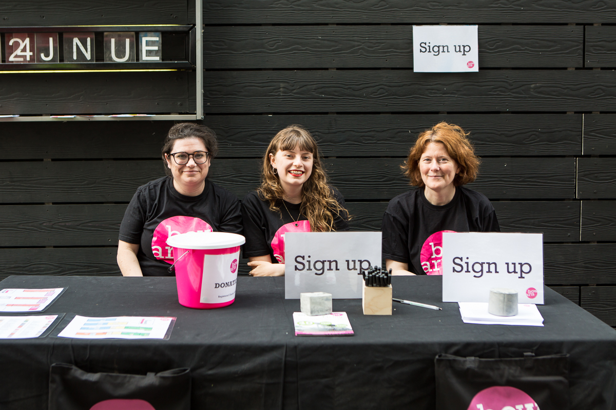 Three Volunteers sit at a welcome desk smiling at the camera