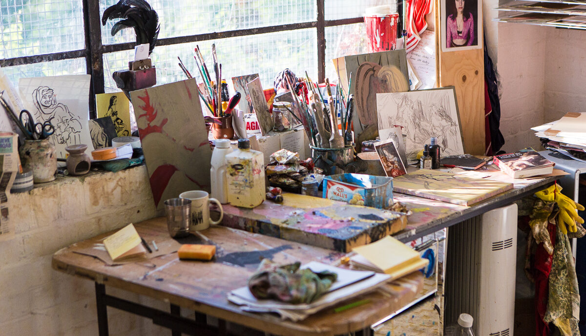 A paint stained desk with paintbrushes, works in progress and other artist tools, in front of a window