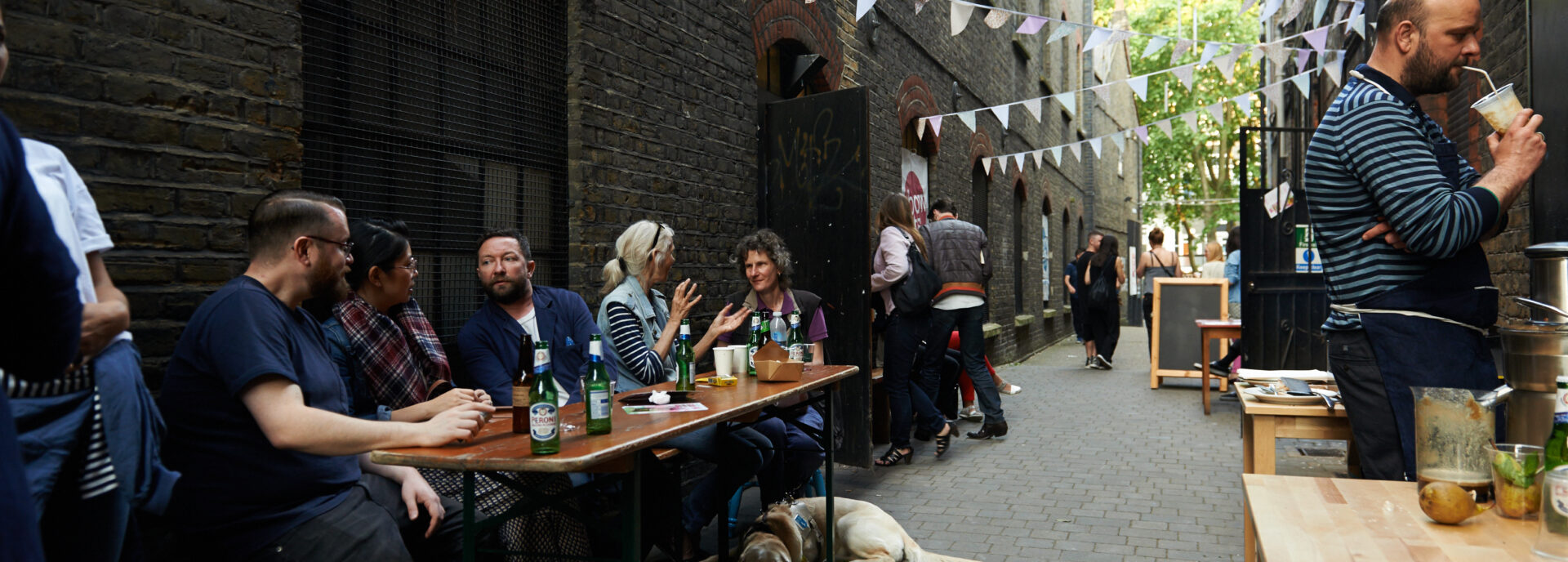 People sit at a table with food and drink in an allyway, pastel bunting is overhead