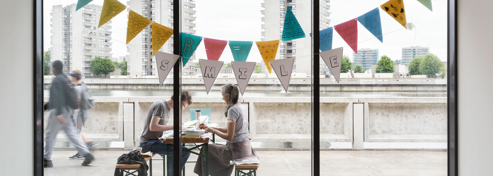 View out of the Lakeside Centre, with two people sat painting on a bench outside, in the background are three brutalist flat buildings