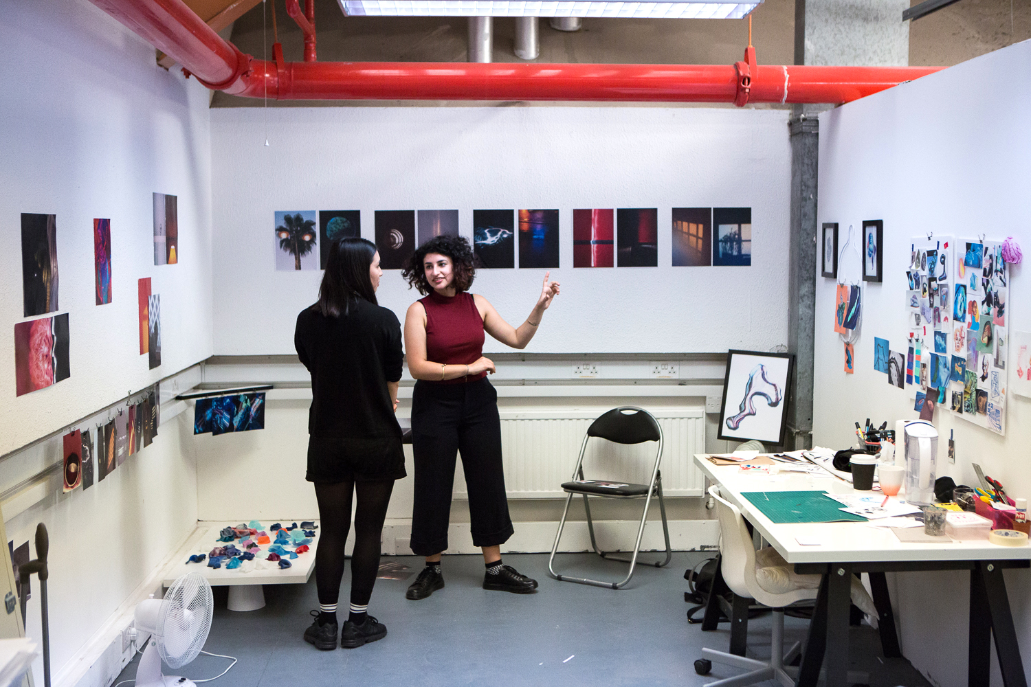 An artist shows someone their work in a studio with photographs on the walls