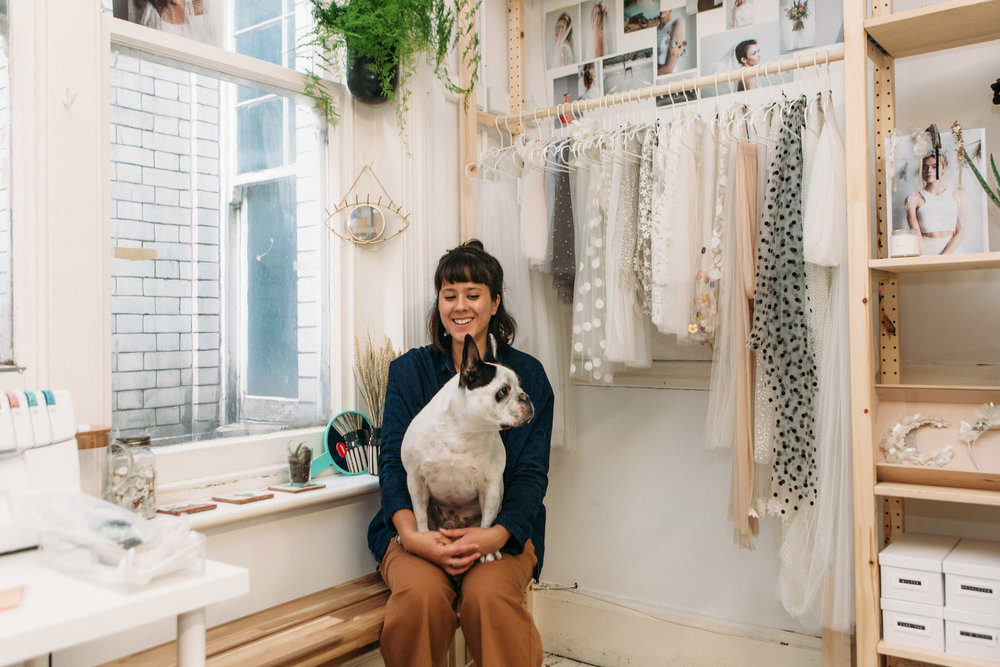 A person sits in their studio with a dog on their lap, behind them white veils hang