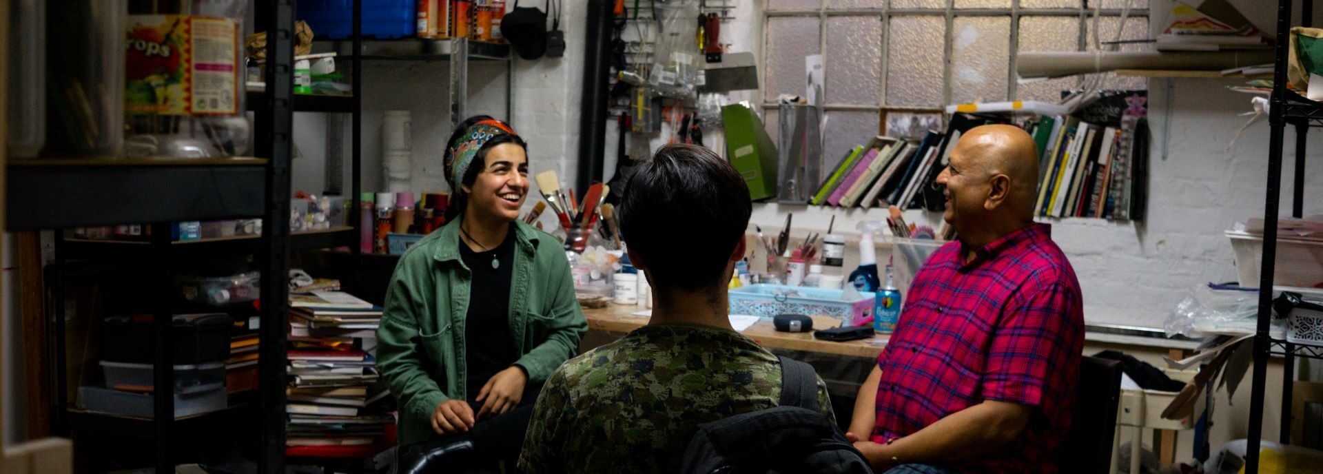 Three people sit in a busy studio, laughing with each other