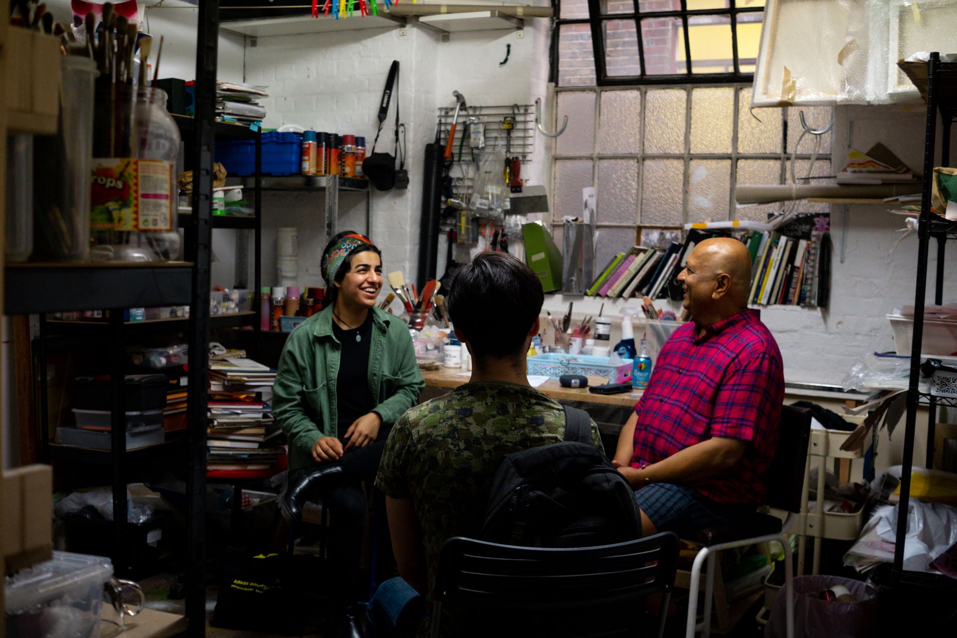 Three people sit in a busy studio, laughing with each other