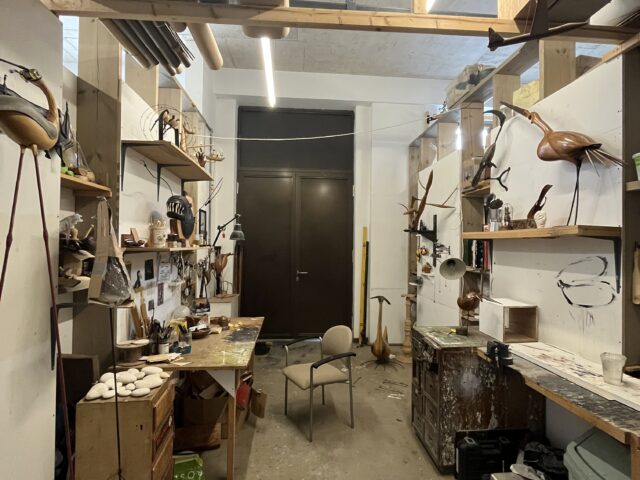 Images shows a rectangular studio with shelves on the left and right hand side walls. At the far end of the studio is a door. Side walls do not reach the ceiling. Lights run the length of the studio with wooden beams just underneath.