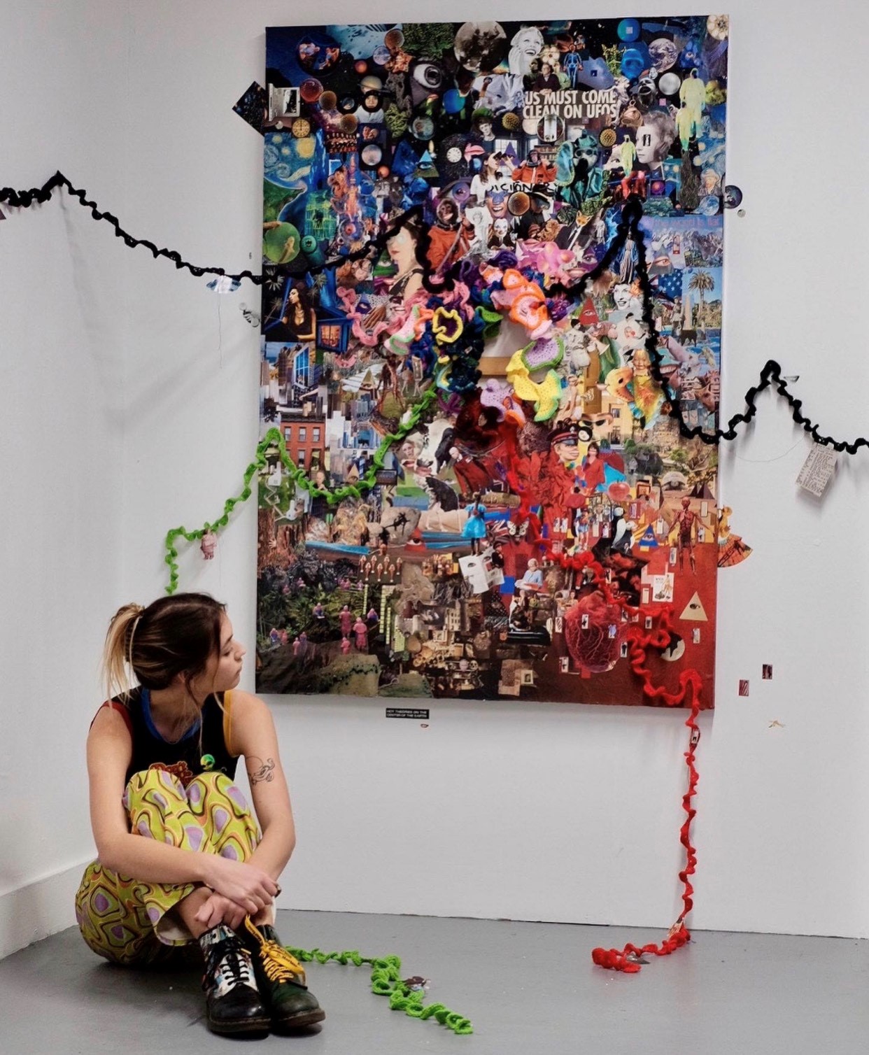 a young white woman sits in front of a chaotic blue, green, and red multimedia artwork