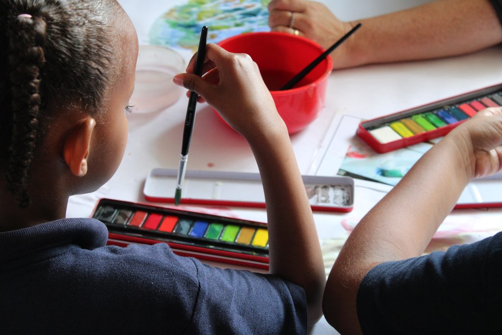 Over the shoulder shot of two children painting with brushes, paint pallet and a container of water